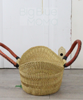 Woven basket with red handles on a wooden floor against a white paneled wall.