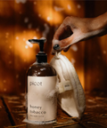 Hand holding a bottle of picot honey tobacco body wash with a towel against a warm, blurred background.
