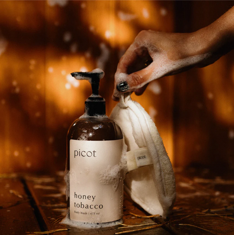 Hand holding a bottle of picot honey tobacco body wash with a towel against a warm, blurred background.