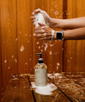 Person washing hands with soap on a wooden surface, with a bottle of soap in the foreground.