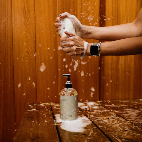 Person washing hands with soap on a wooden surface, with a bottle of soap in the foreground.