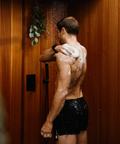 Man showering with soap lather on his back in a wooden shower stall.