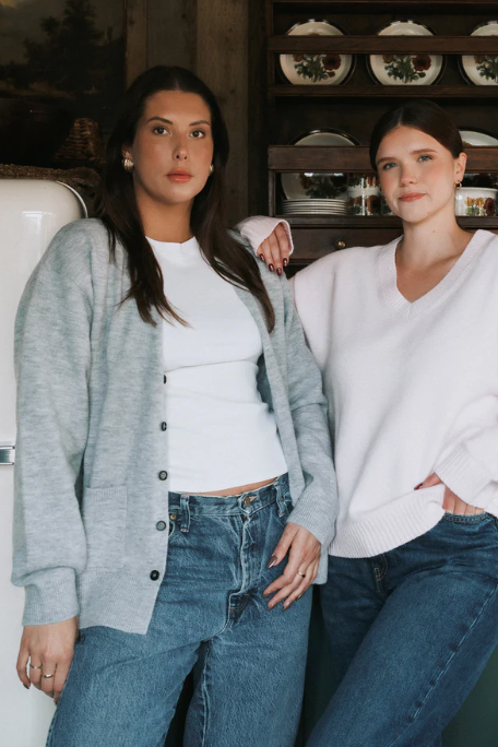Two women standing close together in a casual setting with a shelf in the background.
