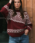 Woman wearing a patterned sweater and jeans standing against a wooden wall.
