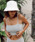 Woman wearing a light gray tank top and white bucket hat outdoors with greenery in the background