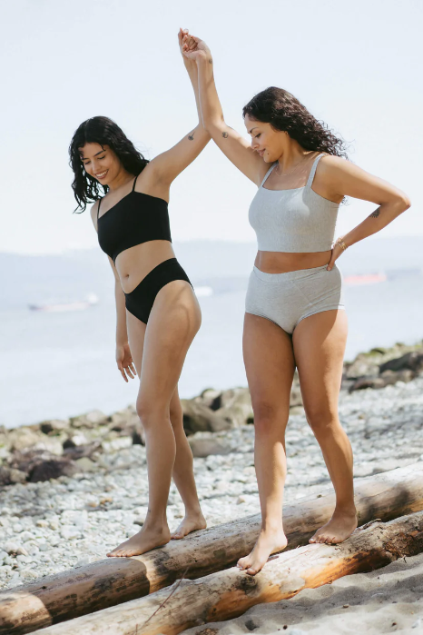 Two women in swimsuits standing on a log by the ocean.