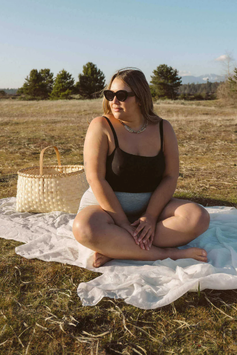 Woman sitting on a white blanket in a field with a woven basket beside her.