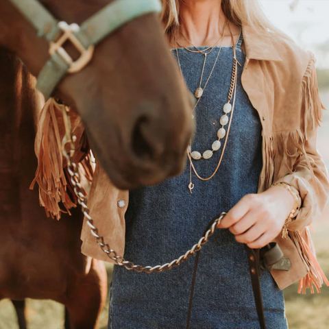 Woman in a denim dress and beige jacket standing next to a horse in an outdoor setting