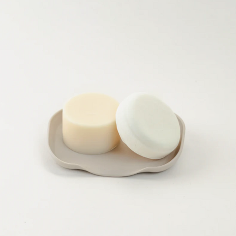 Two white soap bars on a beige soap dish against a light gray background