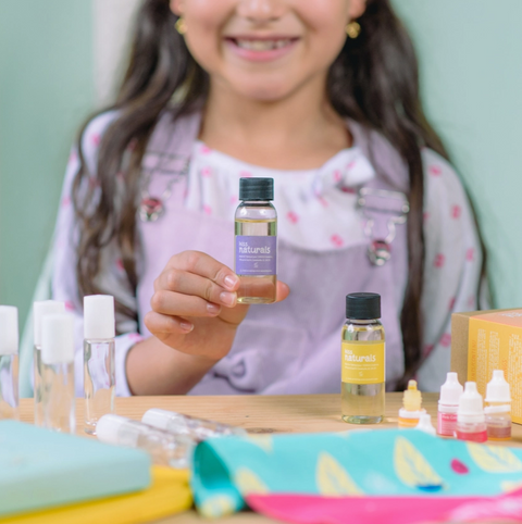 Child holding a bottle labeled 'naturals' with various other bottles and items on a table.