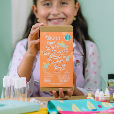 Child holding a 'Miss Naturals' lava lip gloss kit with various lip gloss bottles on a table.
