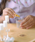 Person pouring a yellow liquid from a small bottle into a clear container on a wooden table.