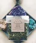 Packaging of Serenity Now calming and anxiety tea by Pachamama. The package is adorned with crystals, has a label and includes a clear window showing the tea leaves inside.
