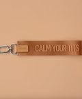 Brown woven keychain with 'CALM YOUR Tits' text on a beige background