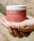 Hand holding a candle labeled 'Raven Song' with a blurred natural background