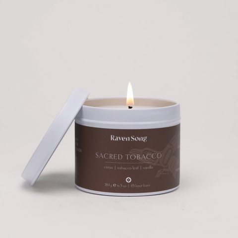 Candle in a brown container labeled 'Raven Song Sacred Tobacco' on a light gray background