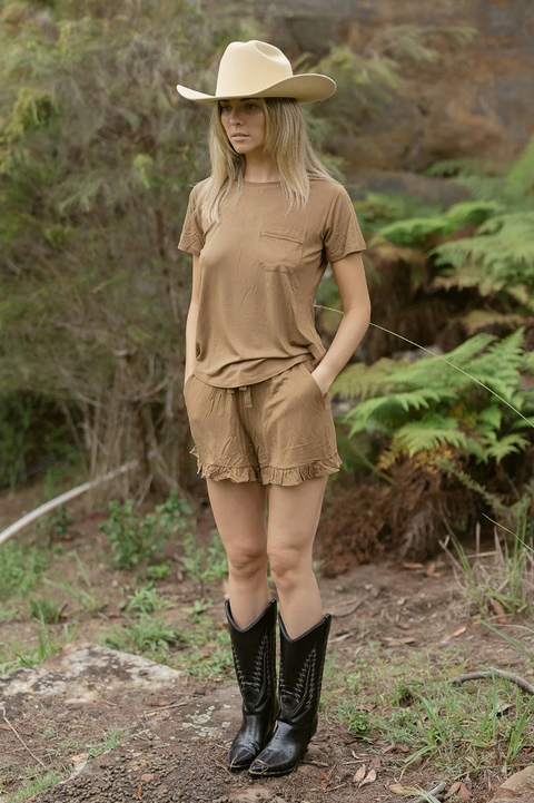 Person wearing a beige cowboy hat, brown shirt, and shorts standing in a natural setting.