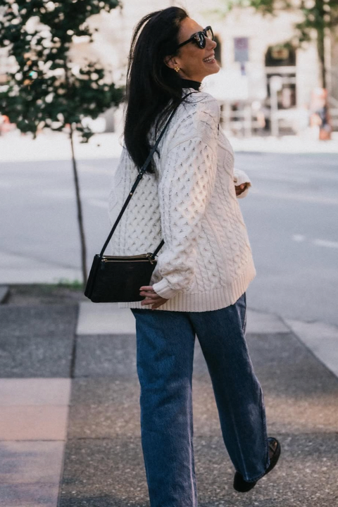 Woman wearing a white cable knit sweater and blue jeans walking on a city street.
