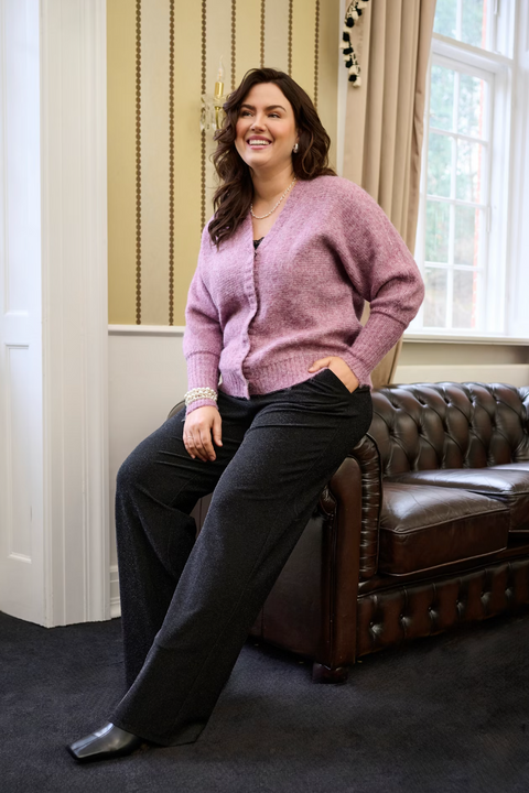 Woman sitting on a leather couch wearing a pink cardigan and dark pants in a room with a window and curtains.