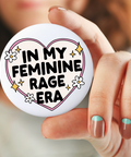 Button badge with 'In my feminine rage era' text held by a hand.