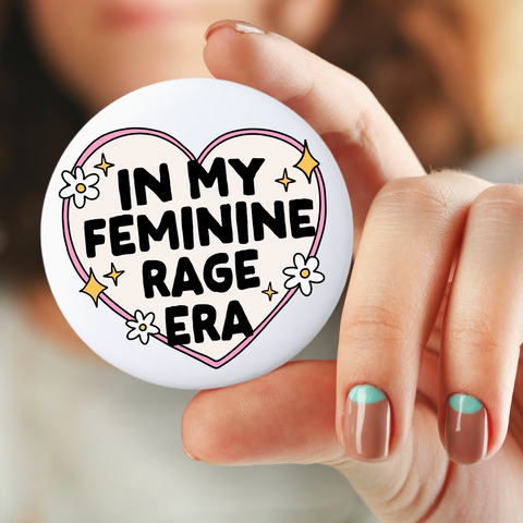 Button badge with 'In my feminine rage era' text held by a hand.