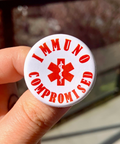 Button badge with 'IMMUNO COMPROMISED' text and medical symbol held by a hand.