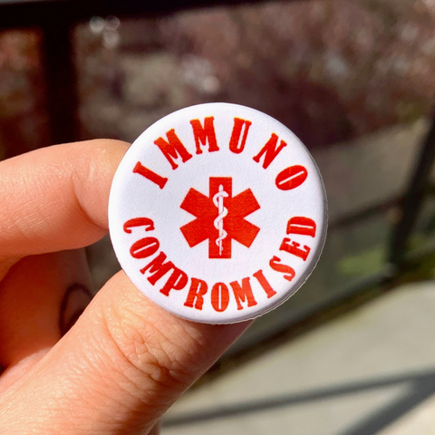 Button badge with 'IMMUNO COMPROMISED' text and medical symbol held by a hand.