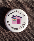 Button badge with 'Housing is a human right' text and house illustration on a textured surface.