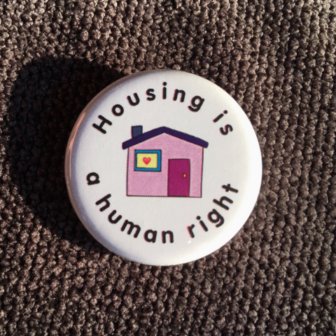 Button badge with 'Housing is a human right' text and house illustration on a textured surface.