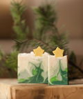 Two green and white soap bars with star-shaped toppers on a wooden surface, blurred greenery in the background.