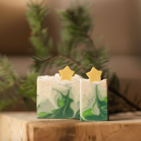 Two green and white soap bars with star-shaped toppers on a wooden surface, blurred greenery in the background.