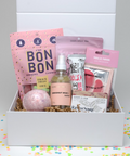 Gift box with various items including a pink bath bomb, gummies, and other products on a colorful confetti background.