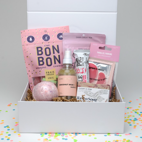 Gift box with various items including a pink bath bomb, gummies, and other products on a colorful confetti background.