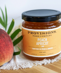 Jar of Provisions Food Company Peach Apricot Jam with a peach and leaves on a wooden surface.