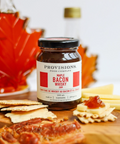 Jar of Provisions Bacon Jam on a wooden board with crackers and bacon, surrounded by other food items.