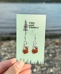 Earring card with 'Tide and Timber' branding held by a hand against a beach background