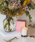 Candle with a white label next to a vase of flowers and pink boxes on a white surface.
