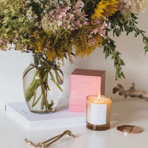 Candle with a white label next to a vase of flowers and pink boxes on a white surface.