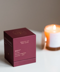 Maroon box labeled 'Poetry of the Gods' next to a lit candle on a white surface.