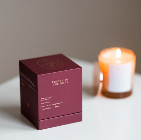 Maroon box labeled 'Poetry of the Gods' next to a lit candle on a white surface.