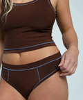 Woman wearing a brown tank top and high-waisted underwear with blue accents on a light gray background.