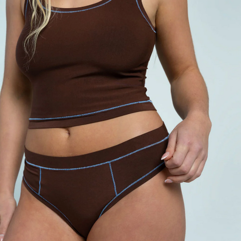 Woman wearing a brown tank top and high-waisted underwear with blue accents on a light gray background.