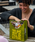 Woman holding a green tote bag with 'This Bag Might Be Full of Kittens' text, sitting at a table outdoors.
