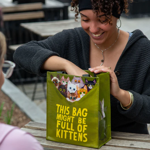 Woman holding a green tote bag with 'This Bag Might Be Full of Kittens' text, sitting at a table outdoors.