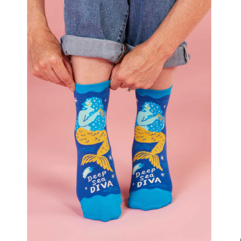 Person wearing blue socks with mermaid design and 'Deep Sea Diva' text on a pink background.