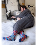 Man sitting on a bed wearing colorful socks with a laptop, surrounded by cat-themed products.