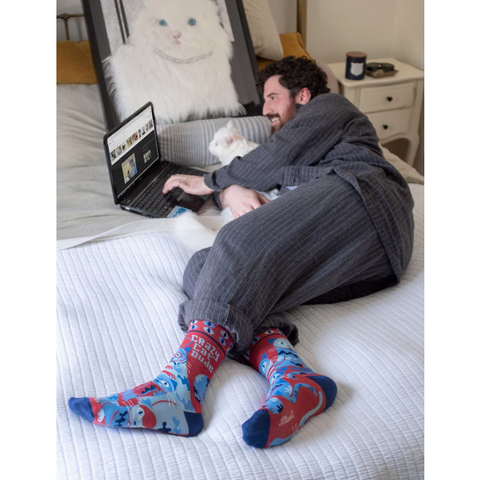 Man sitting on a bed wearing colorful socks with a laptop, surrounded by cat-themed products.