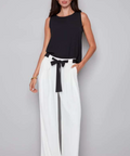 Woman wearing a black sleeveless top and white wide-leg pants with a belt against a gray background