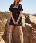 Woman in a black polka dot dress and beige boots standing on rocks with a clear sky background