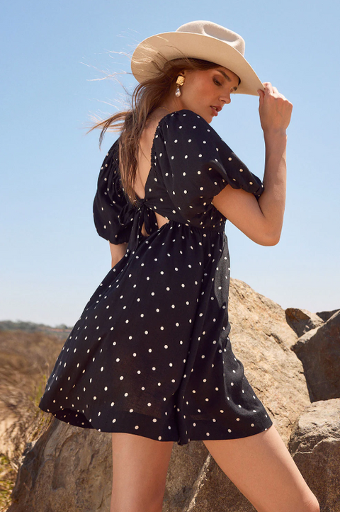 Woman in a black polka dot dress and beige cowboy hat standing on rocky terrain with a clear blue sky.